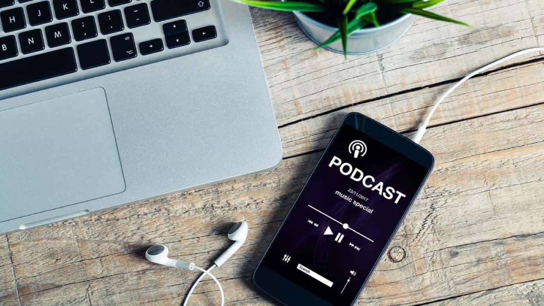 Dizüstü bilgisayarın yanında duran ve kulaklık takılı olan cep telefonunda açık olan podcast yayını