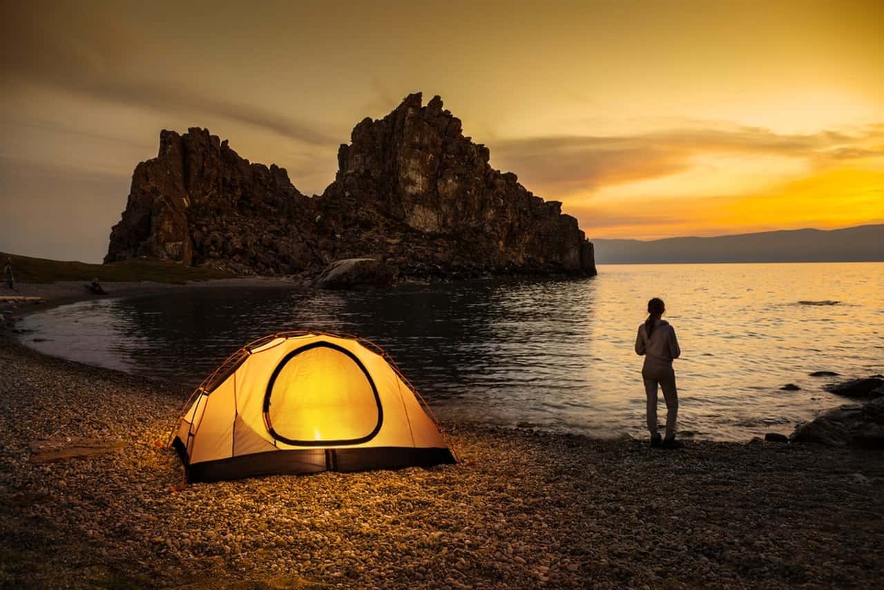 Deniz kenarında gecenin karanlığında içindeki aydınlatma görünen çadır ve denize doğru yürüyen arkası dönük kadın