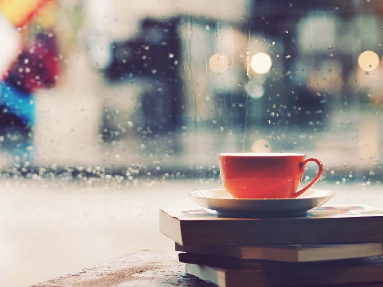 yağmur damlalarının vurduğu camın kenarında üst üste duran kitapların üzerine konmuş bir fincan kahve