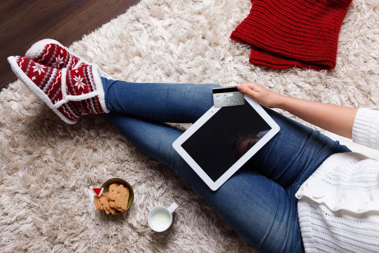 ahşap zemin üzerindeki halıda bacaklarını uzatarak oturan ve tabletten online alışveriş yapan kadın